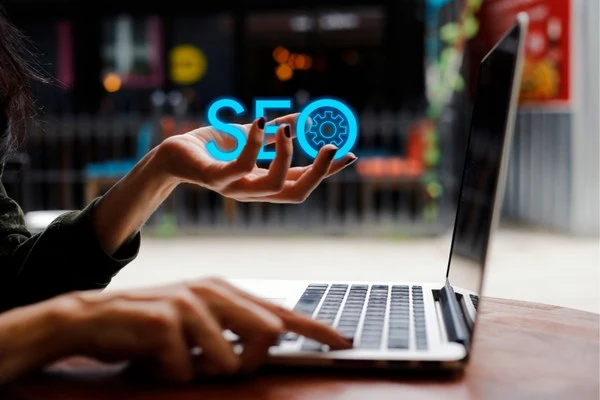 A person using a laptop with the text "SEO" and a gear icon above their hand, representing SEO tools for ranking on Google.