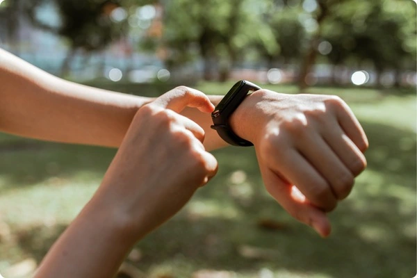 In a sunlit park, a person taps the screen of a black fitness-tracker wearable technology strapped to their wrist.